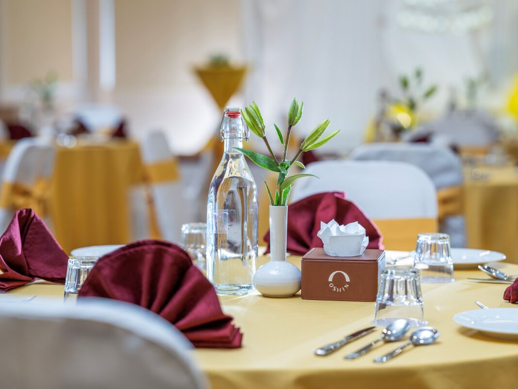 A dining table set with a bottle, a vase with green leaves, a tissue box, and cutlery.