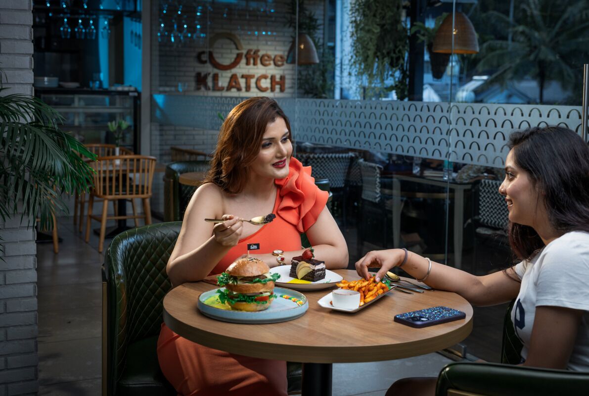 Two people dining at a table in a modern cafe with a visible menu and a sign reading "Coffee Klatch".