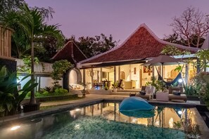 Zona de piscina con un espacio de descanso techado, piscina y vegetación circundante.