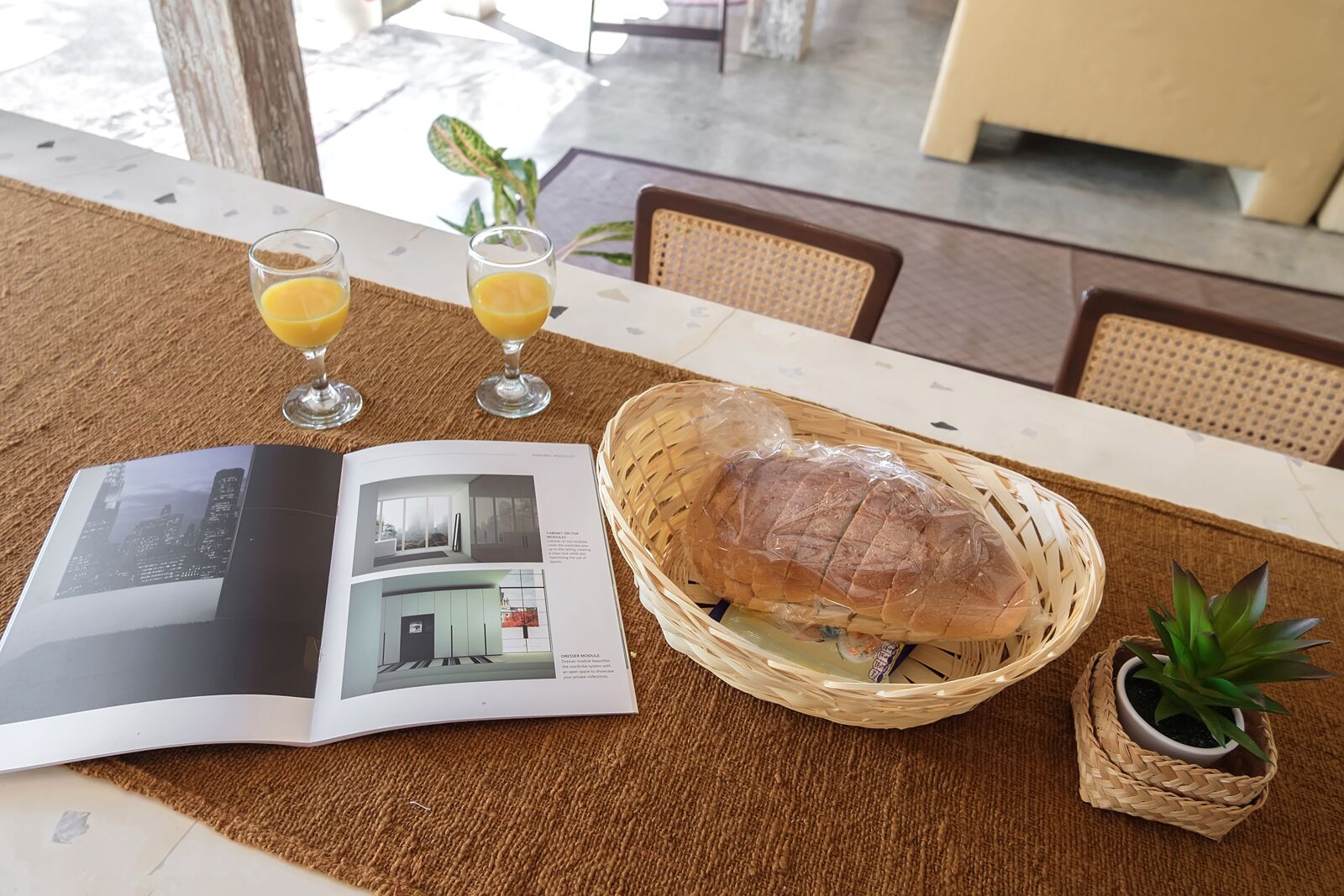 A table with a magazine, two glasses of orange juice, a loaf of bread in a basket, and a small potted plant.