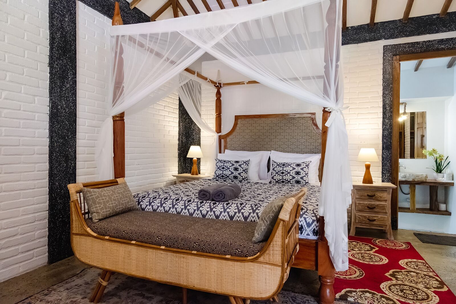 A room with a four-poster bed, a wicker chair, a red patterned rug, and a wooden nightstand with a lamp.