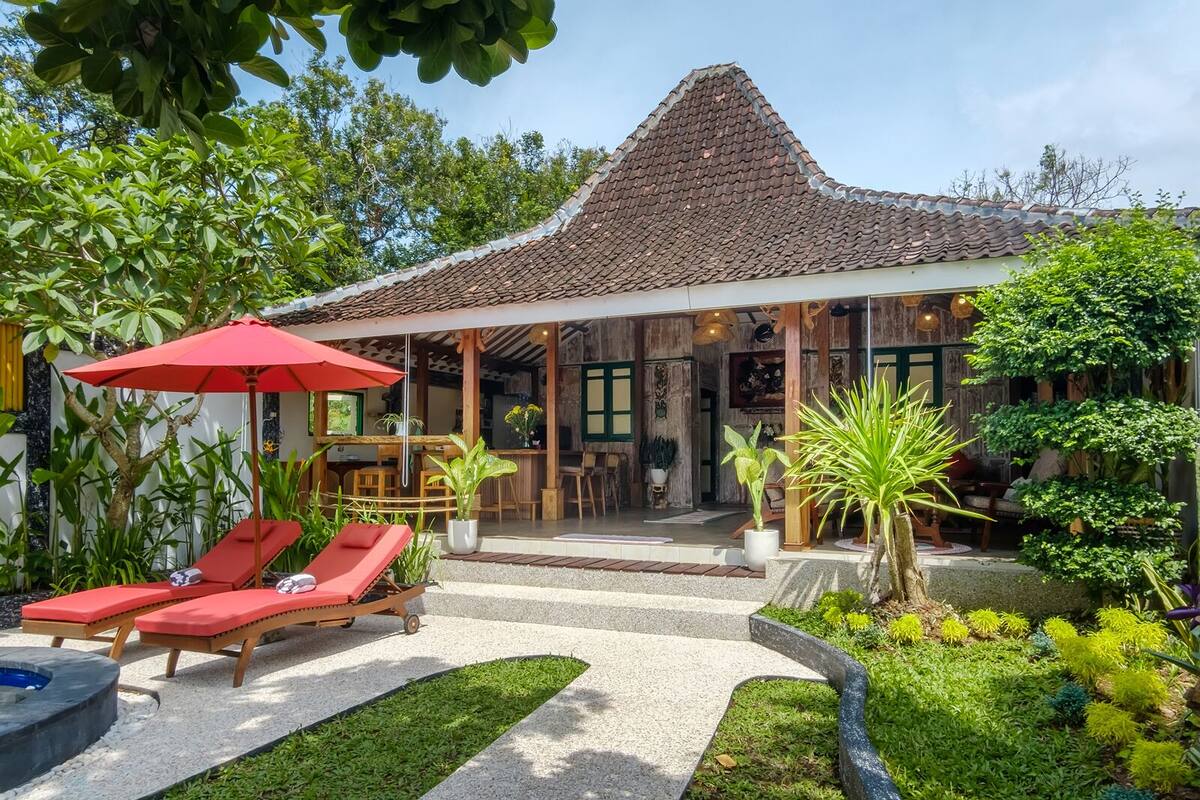 A traditional house with a tiled roof, a covered patio with red lounge chairs, and a garden with various plants.
