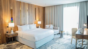 Una habitación de hotel con una cama grande, dos mesitas de noche con lámparas, un área de descanso con una silla y una pequeña mesa, y un televisor sobre un mueble.