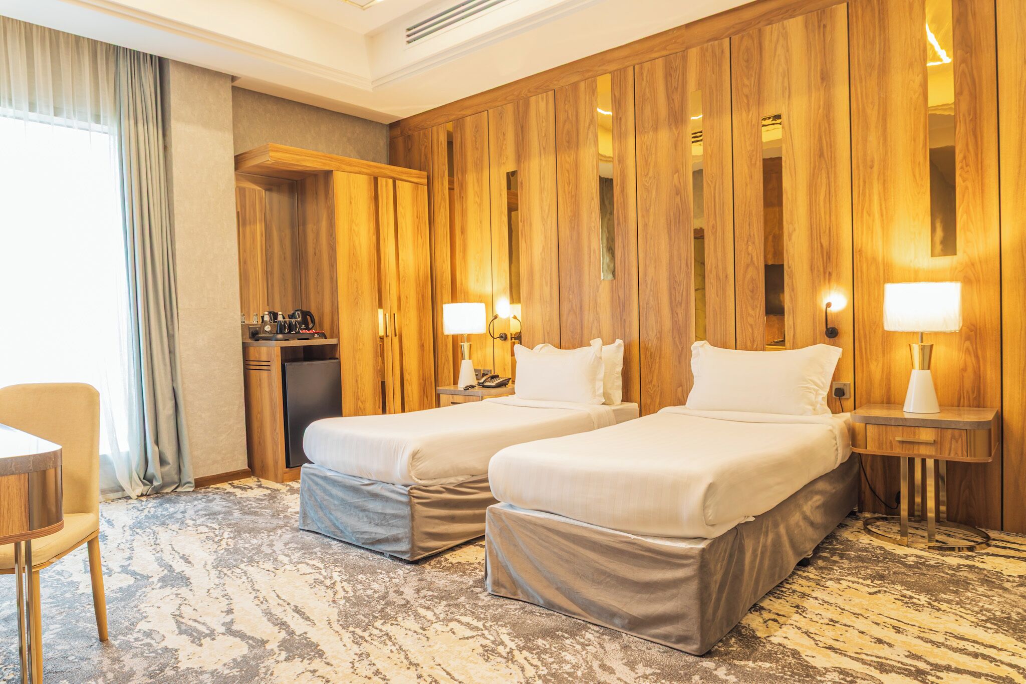 A hotel room with two beds, a desk, a chair, a small kitchenette, and wooden paneling on the walls.