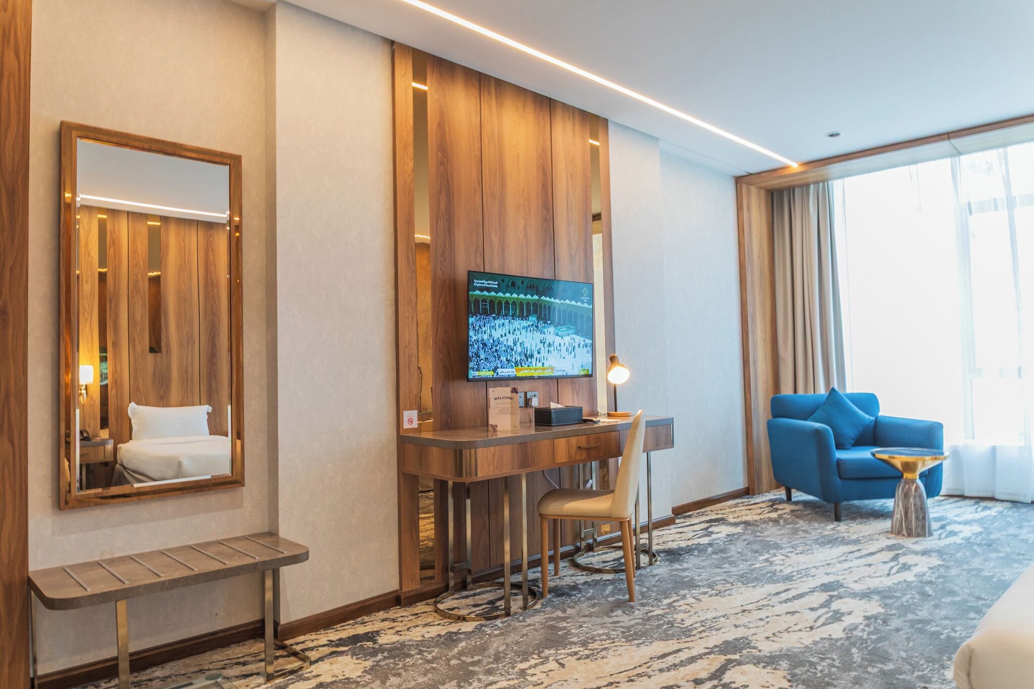 Una habitación de hotel con un escritorio de madera, un televisor, un sillón azul y un banco.