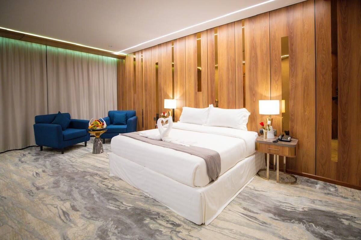 Habitación de hotel con una cama grande, dos sillones azules, una mesita con un florero y paneles de madera en las paredes.