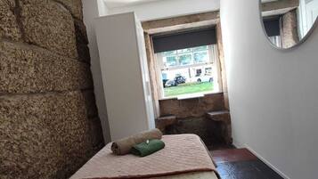 A room with a stone wall, a bed with a patterned bedspread, a small window, and a mirror on the wall.