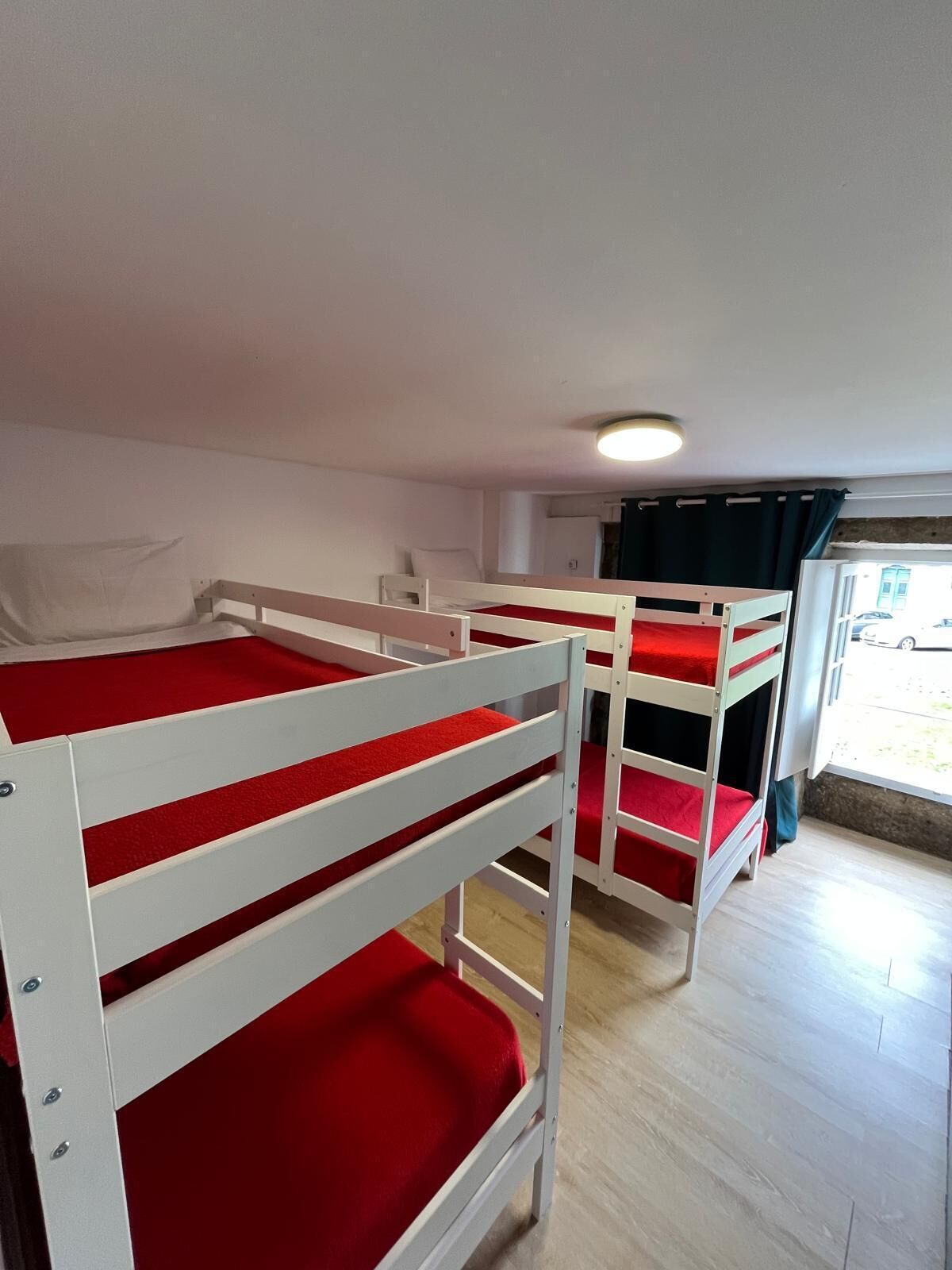 Camas literas con ropa de cama roja en una habitación con piso de madera y una ventana con cortinas.