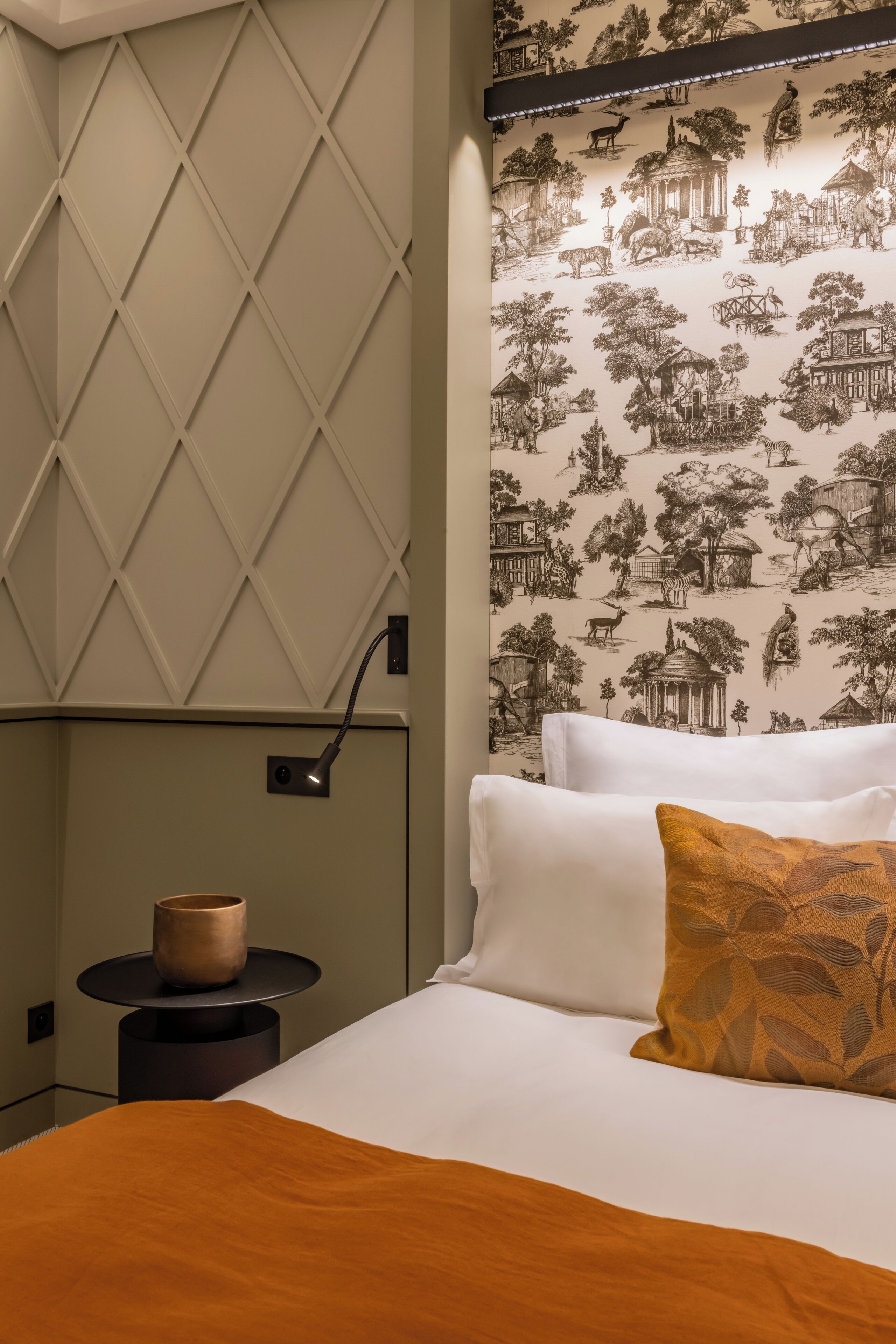 A neatly made bed with white linens and a single decorative pillow, a bedside table with a small vase, and a wall with a geometric pattern and a detailed wallpaper depicting various scenes.