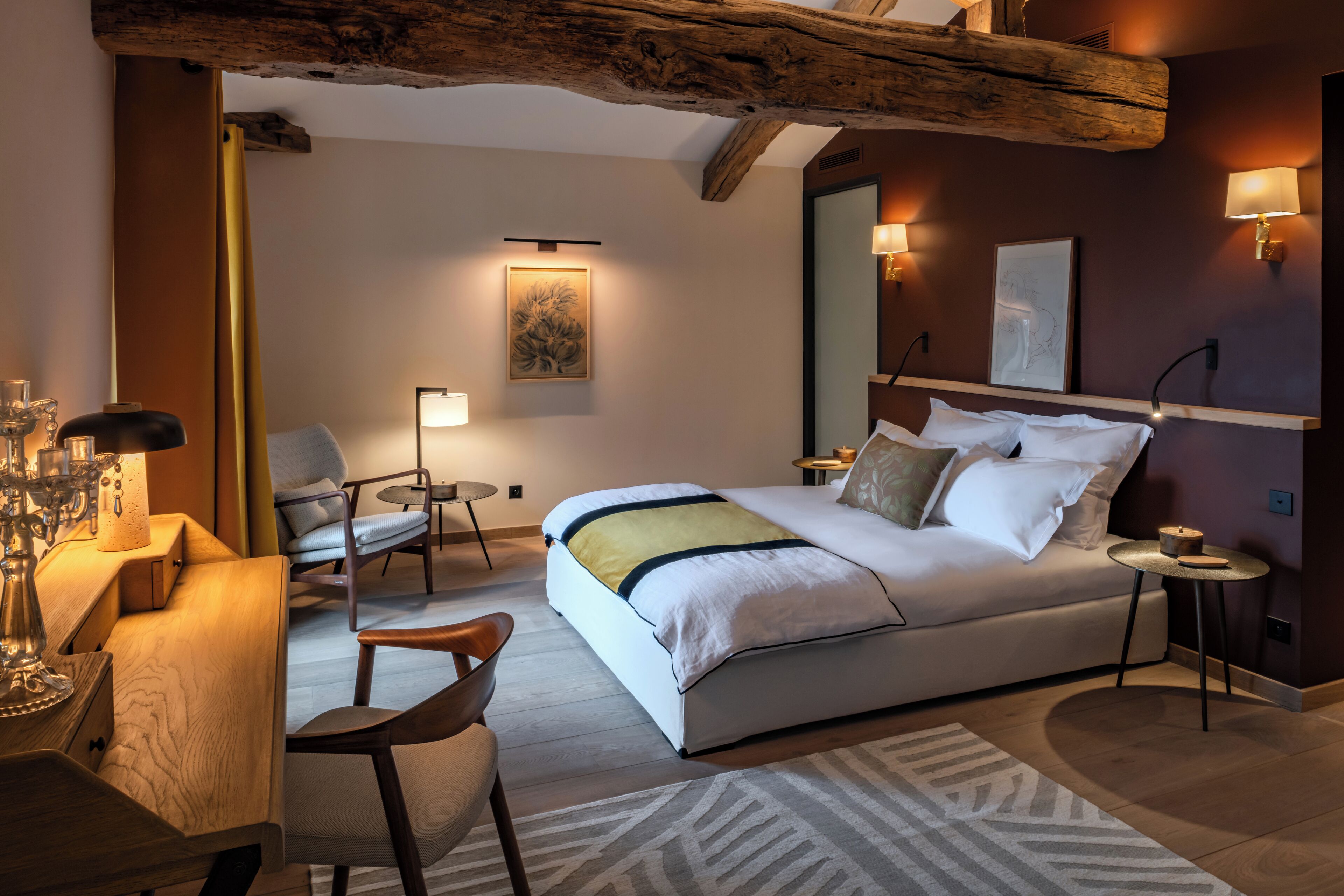 A modern hotel room with a large bed, a desk with a lamp, a chair, and a small round table.