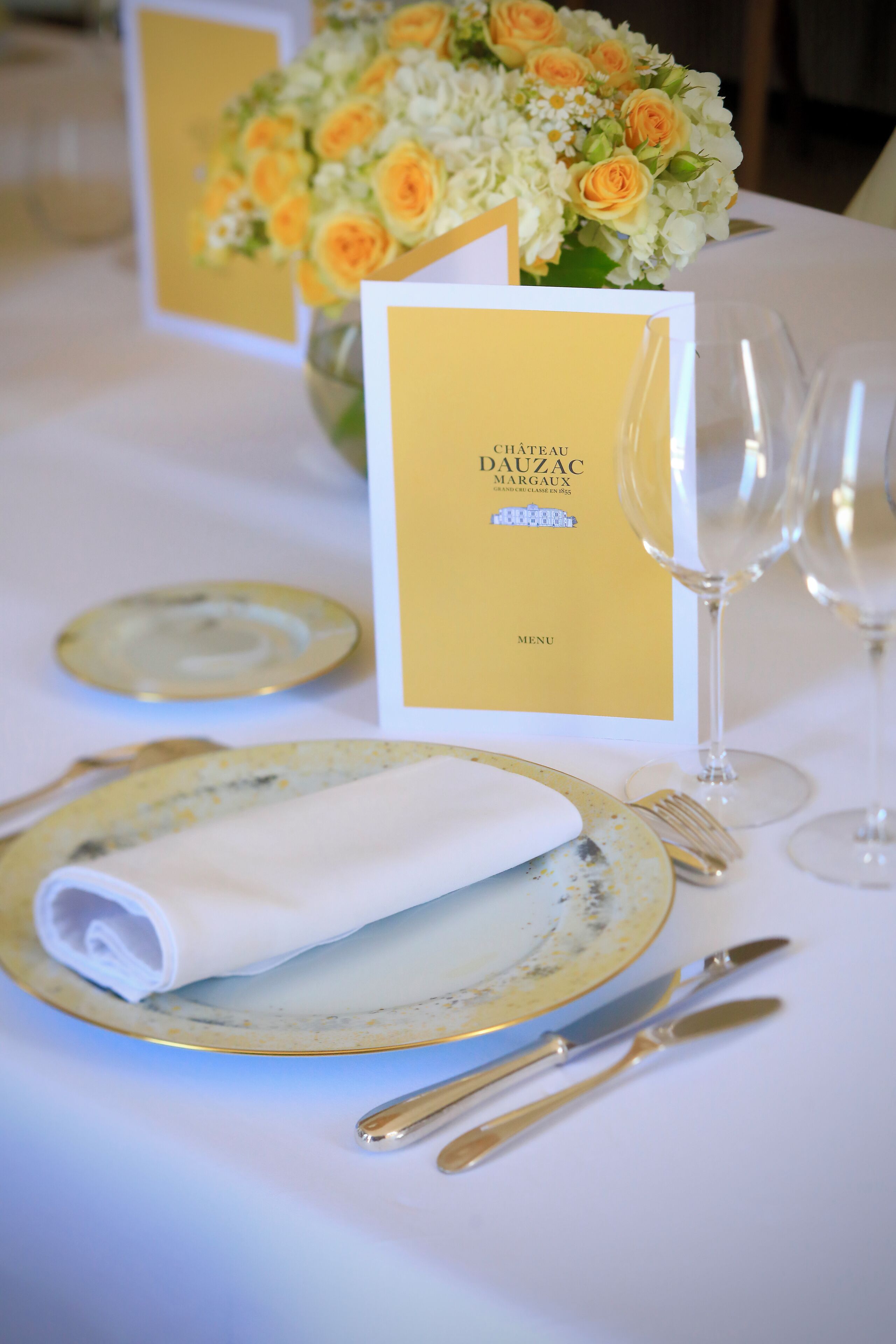 Ein Tisch ist mit goldenen und weißen Tellern, Besteck und einem gefalteten Servietttuch gedeckt. Eine Speisekarte und eine Vase mit Blumen sind ebenfalls zu sehen.
