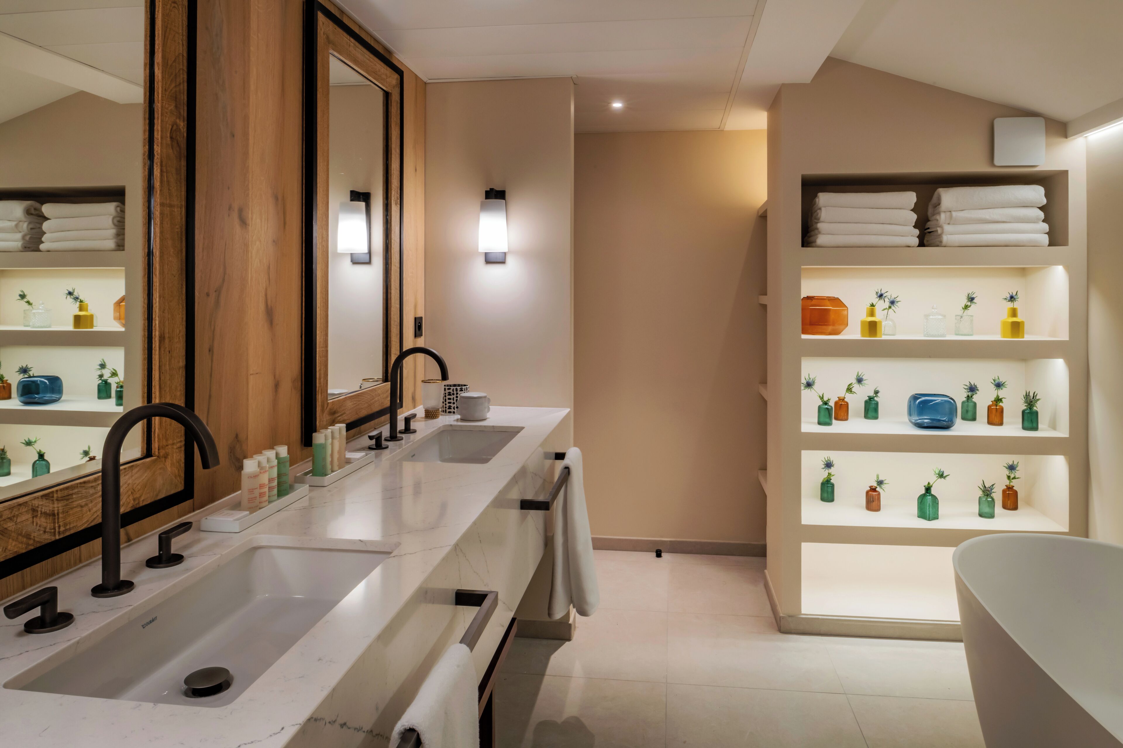 A modern bathroom with a large mirror, two sinks, a bathtub, and shelves with toiletries.