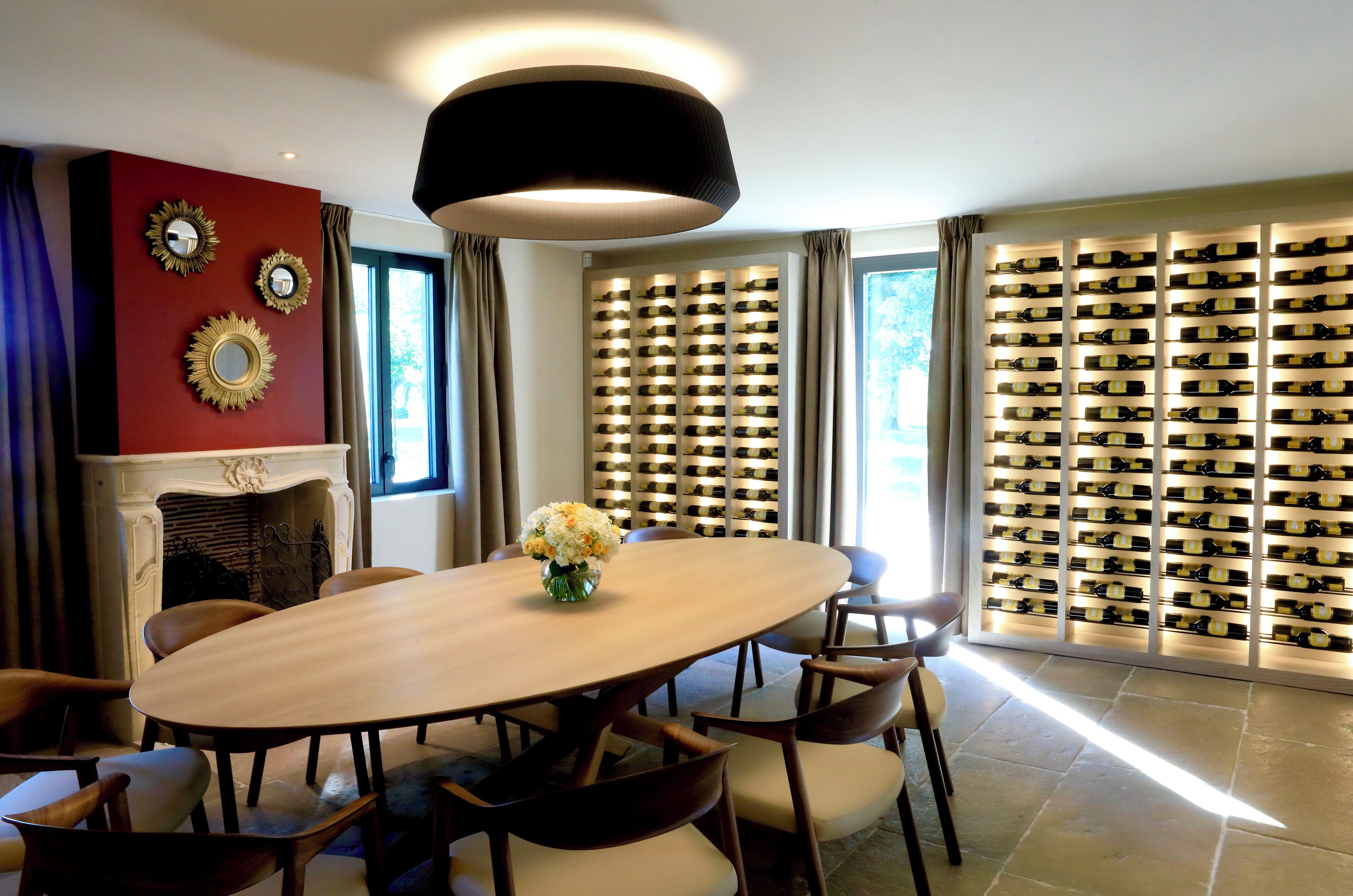 A dining area with a large oval table, chairs, a vase with flowers, a fireplace, and a wine rack.