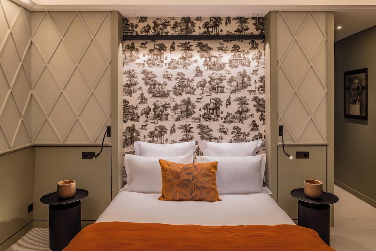 Un dormitorio ordenado con un papel tapiz estampado, una cama con ropa de cama blanca y marrón, y dos mesitas de noche con pequeños jarrones.