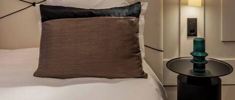 A modern hotel room with a neatly made bed, two framed abstract artworks on the wall, a black bedside table with a teal lamp, and a wall-mounted light fixture.