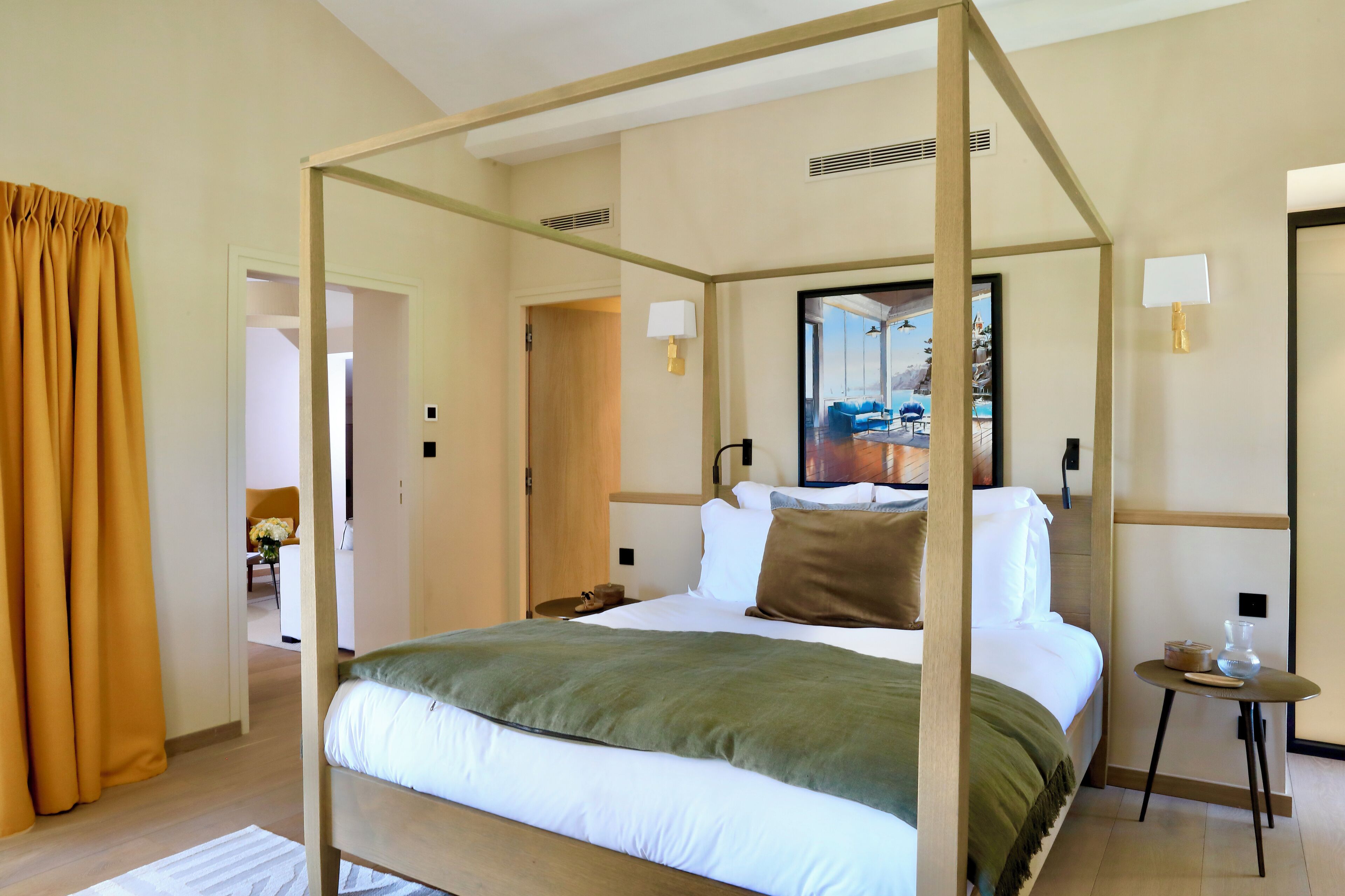 A four-poster bed with a green throw, a bedside table with a lamp, and a view of a beach through a large mirror.