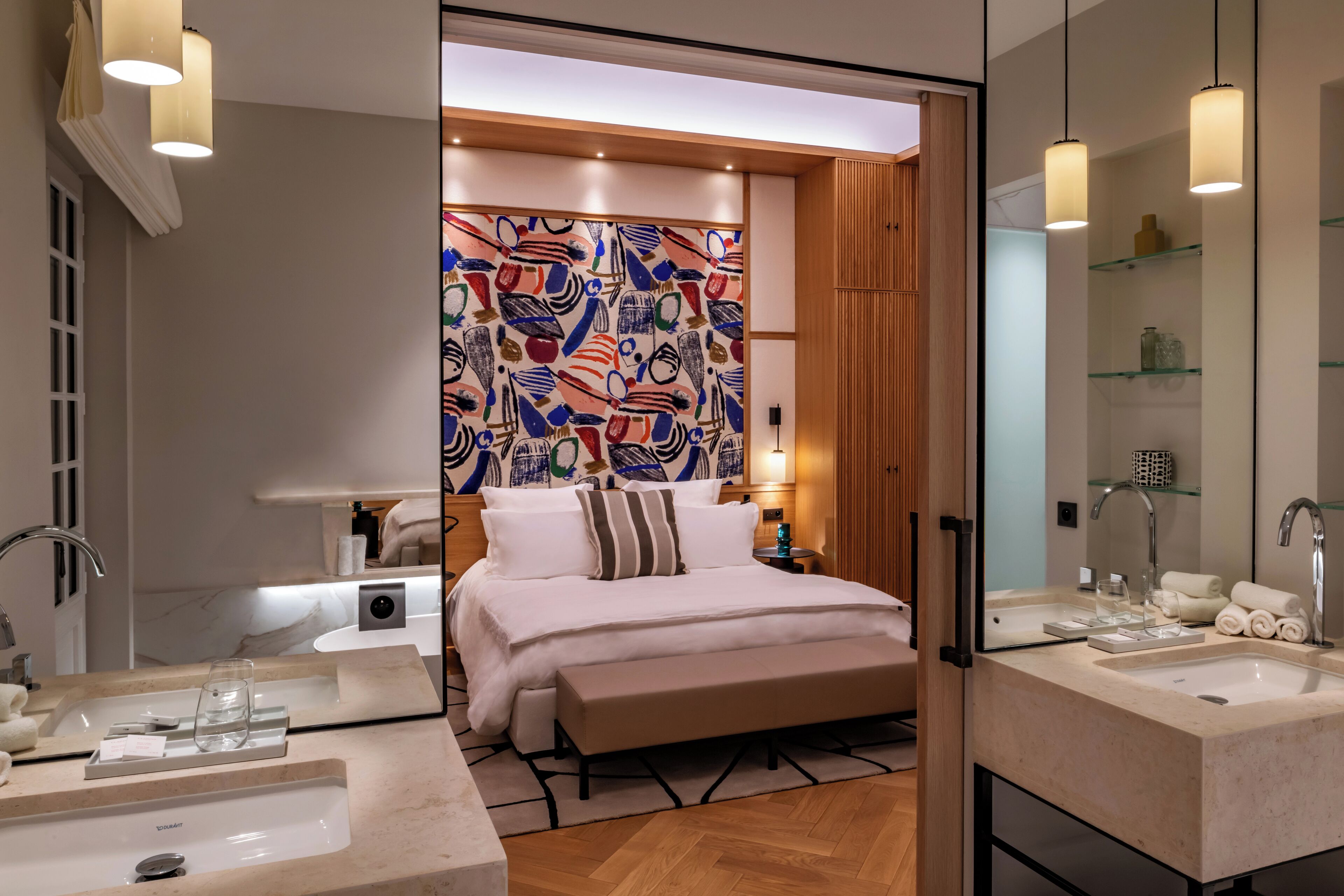 A modern hotel room with a large bed, two bedside tables, a large mirror, a bathroom with a sink and mirror, and a colorful abstract wall art.