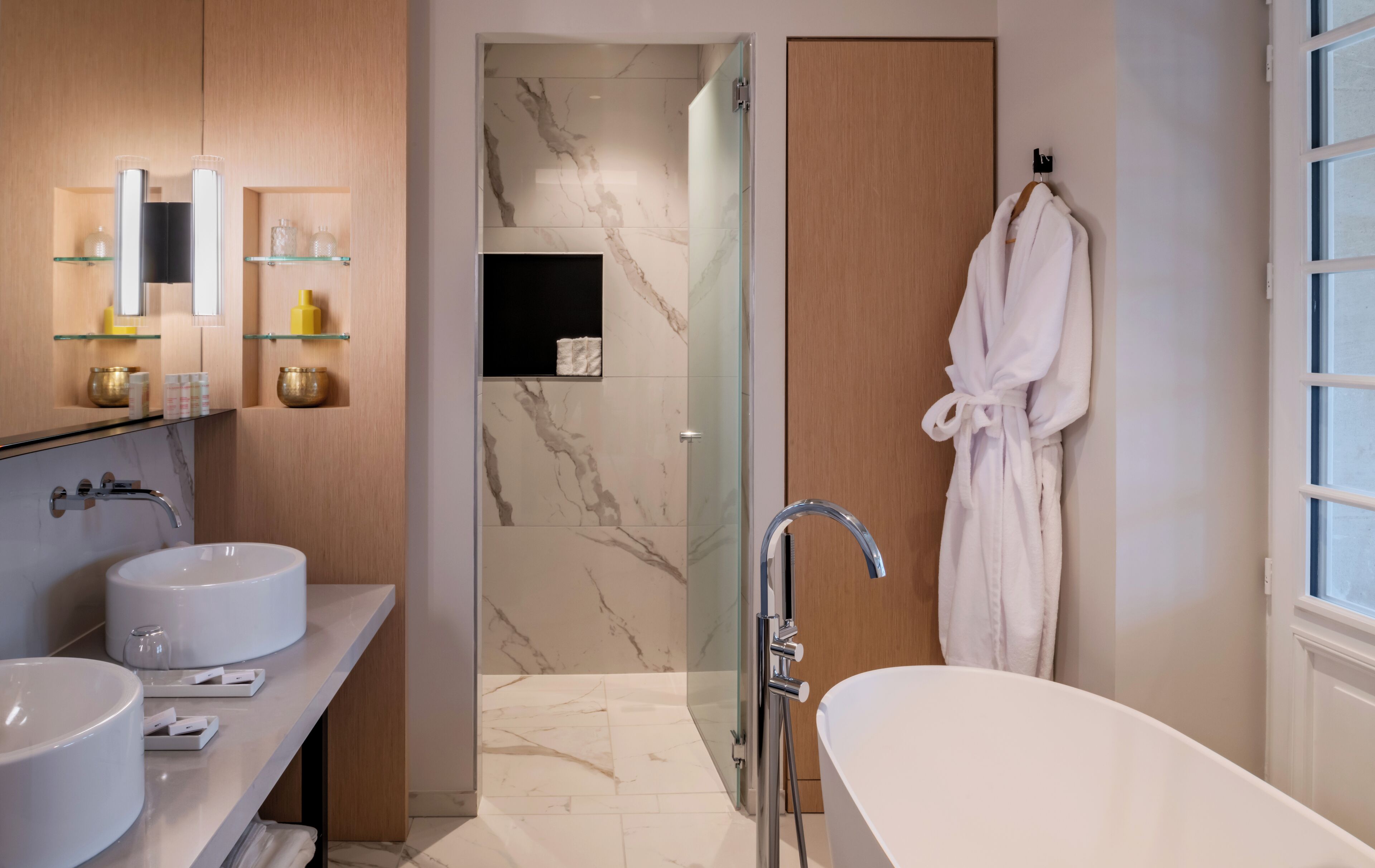 A modern bathroom with two sinks, a glass shower enclosure, a bathtub, and a robe hanging on the wall.