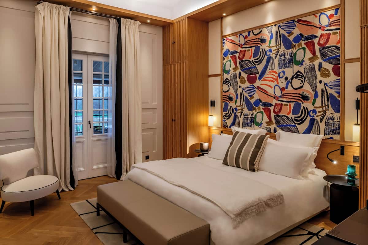 Un dormitorio con una cama grande, una silla, una mesita de noche y un cuadro abstracto y colorido en la pared.