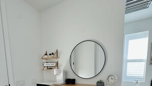 Eine kompakte Küche mit einer Mikrowelle, einem Kühlschrank und einer kleinen Kaffeemaschine. Unter einem Holztisch stehen zwei Hocker, und ein runder Spiegel hängt an der Wand.