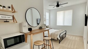 Ein kleines Schlafzimmer mit einem Bett, einem Schreibtisch mit zwei Hocker, einer Mikrowelle, einem Kühlschrank, einem Deckenventilator und einem Fenster mit Jalousien.