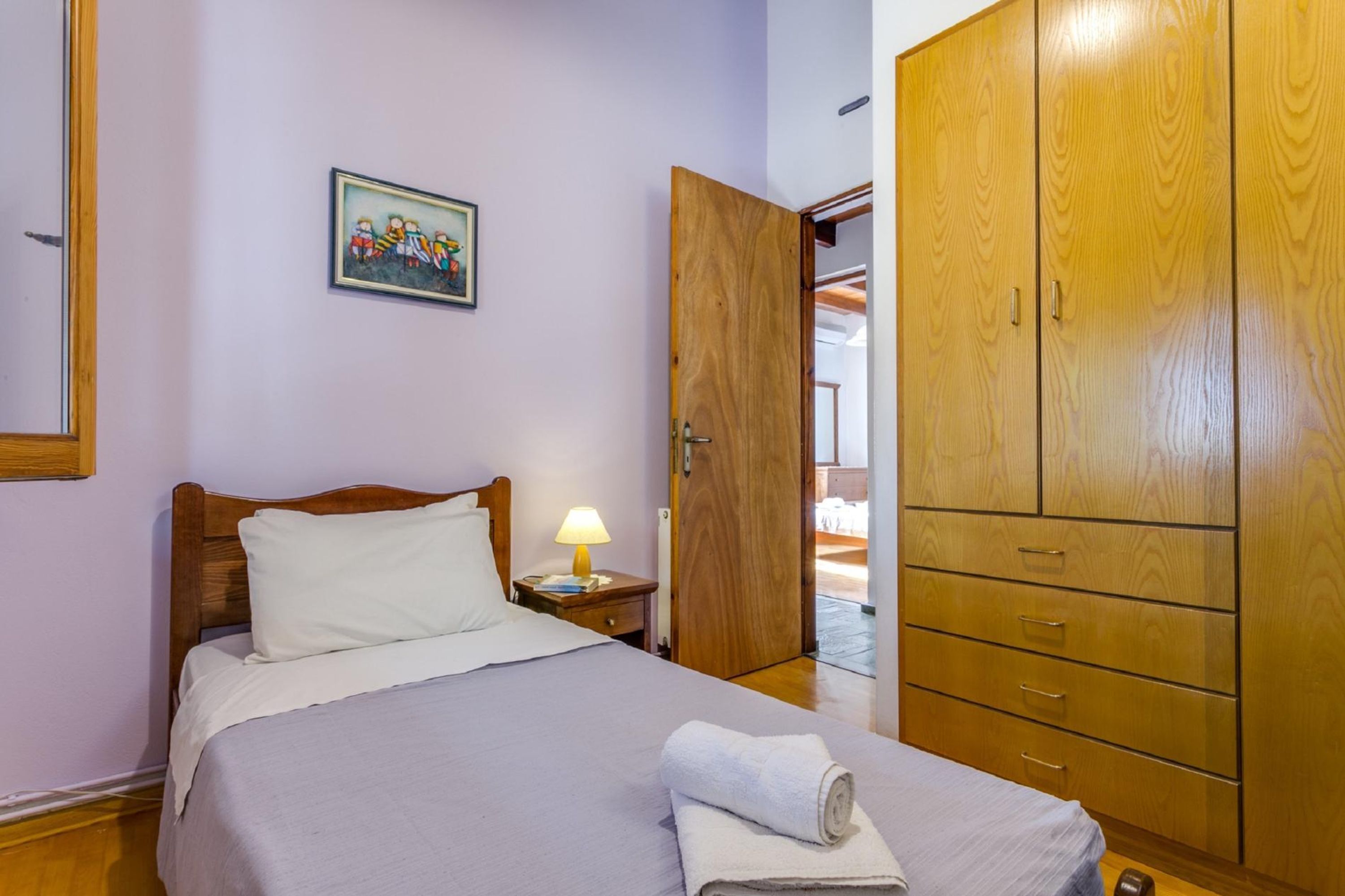Quarto com cama, mesa de cabeceira com luminária, guarda-roupa de madeira e um quadro na parede.
