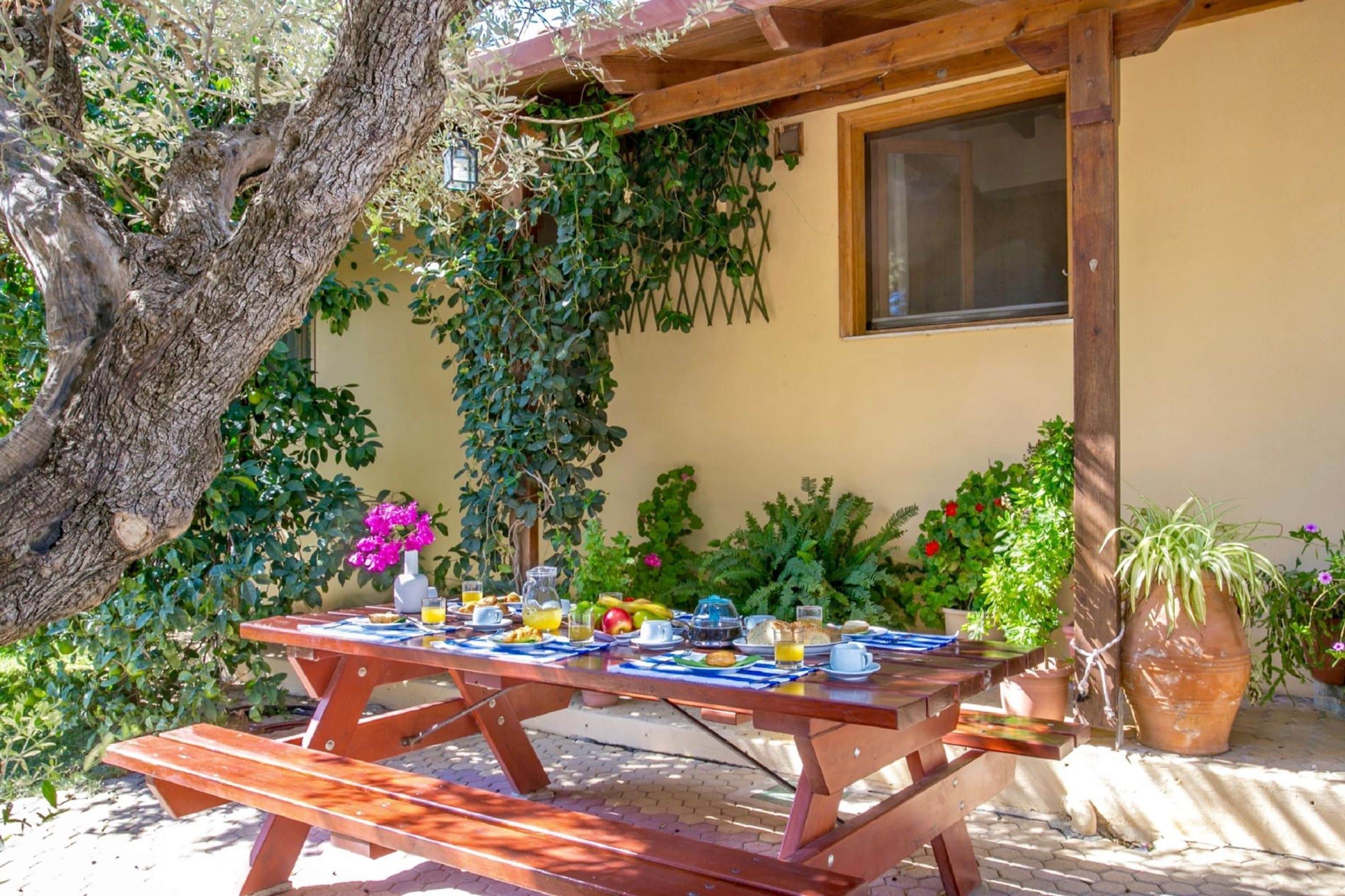 A wooden picnic table set up in a garden with a yellow wall and various plants in the background.