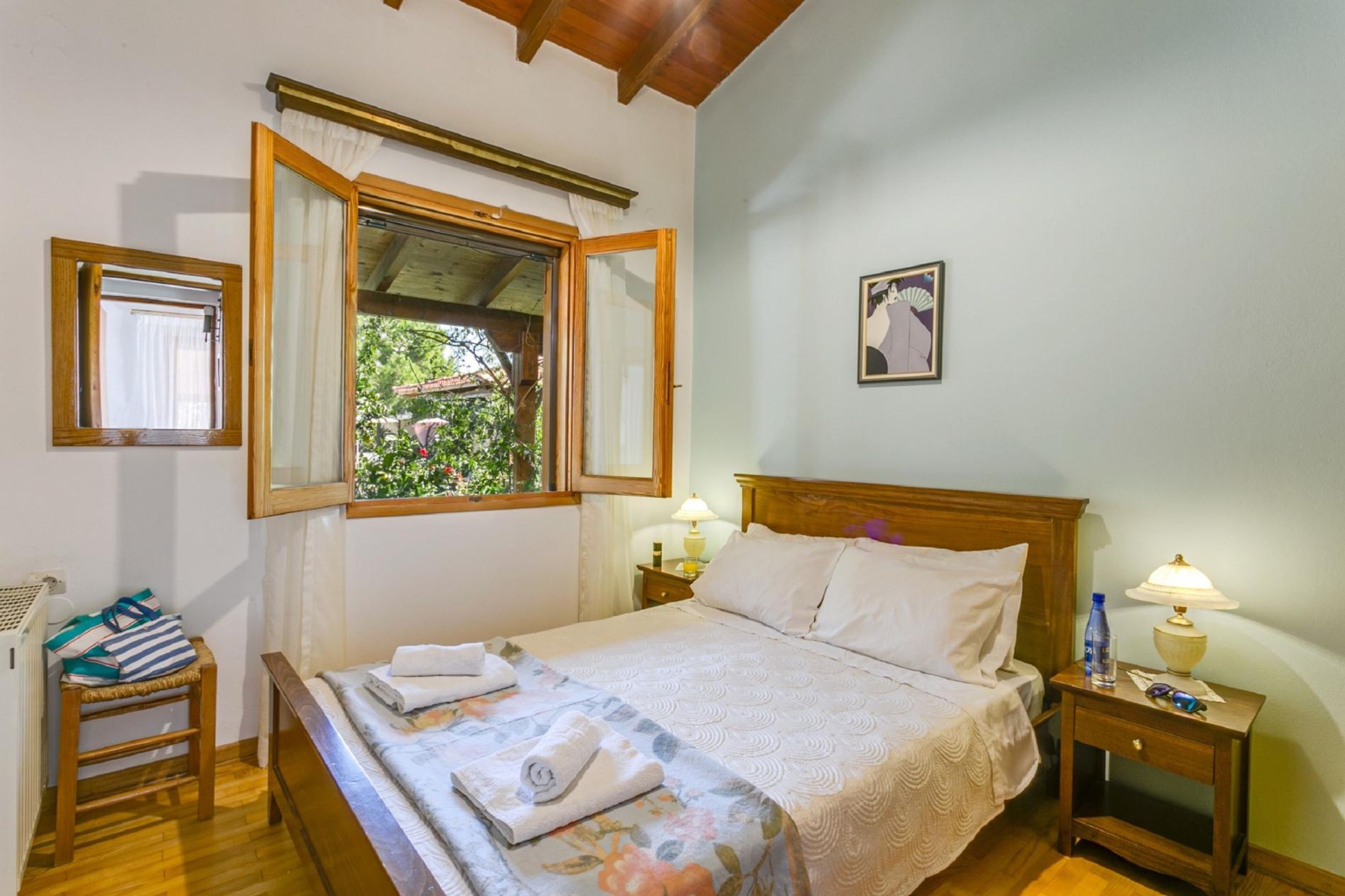 A bedroom with a wooden bed, a nightstand with a lamp, a window with curtains, and a framed picture on the wall.
