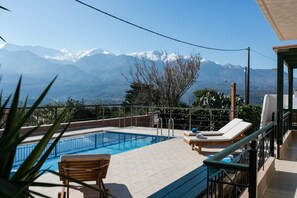 A pool area with a view of snow-capped mountains, a wooden deck, and a lounge chair. - Villa Aloni in Chania (Apokoronas)