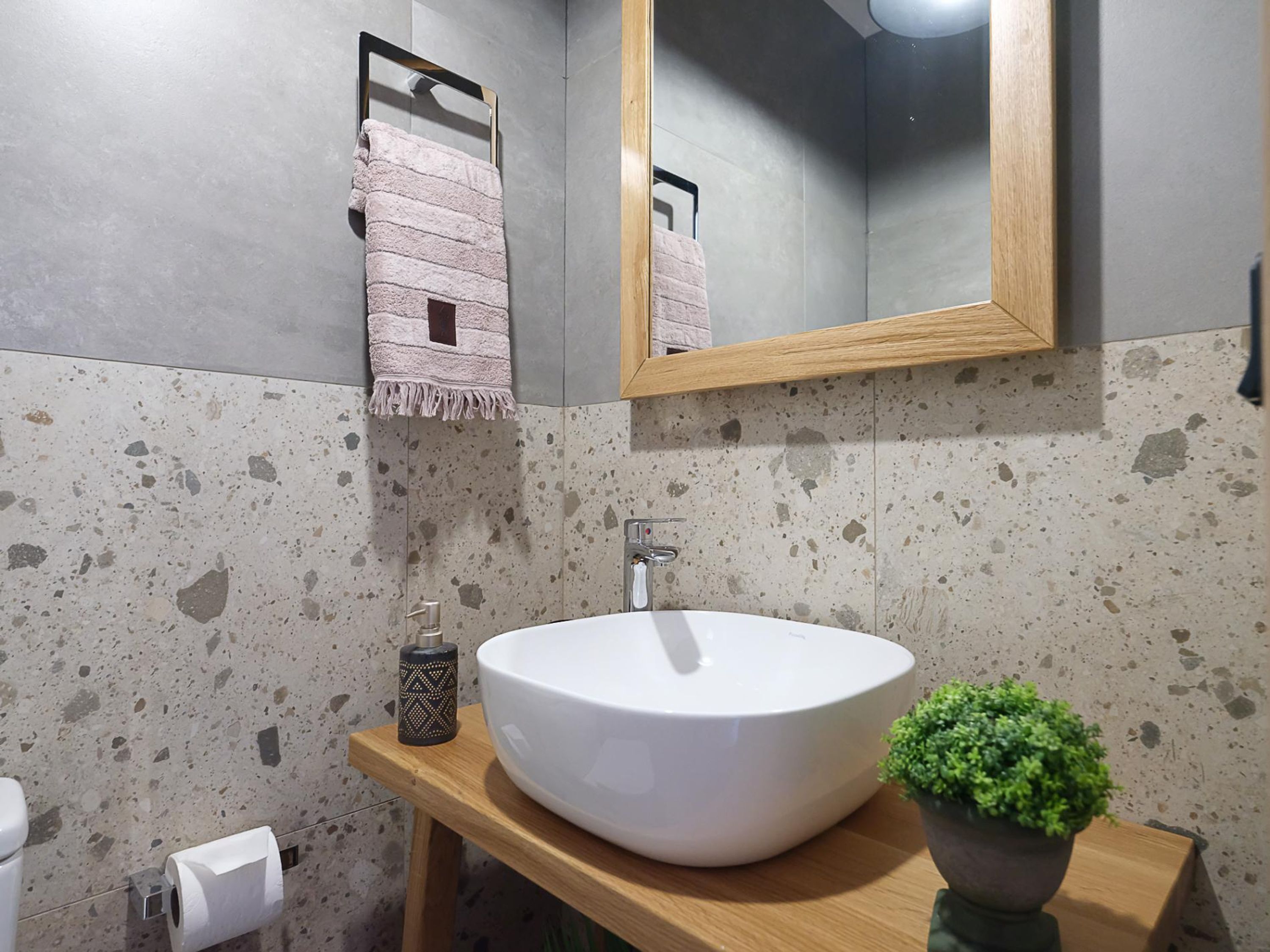 Un baño con un lavamanos grande blanco, un mueble de tocador de madera, un espejo, un perchero para toallas y una pequeña planta en maceta.