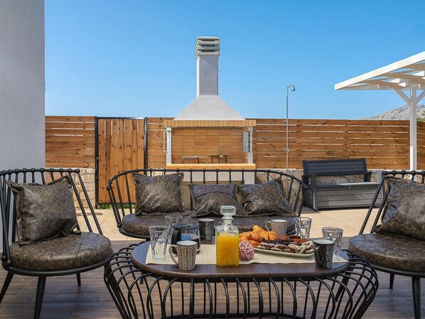 An outdoor patio with a barbecue grill, seating area with chairs and cushions, and a table set with food and drinks.