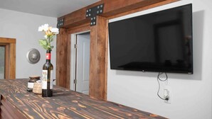 A wooden bar counter with a bottle and flowers, a mounted TV, and a door with a wooden frame. - Meadow Rue Retreat • Aurora Views Near Fairbanks (North Pole, AK)