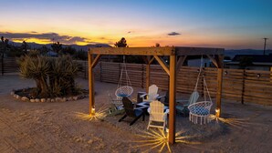 A wooden pergola with swing seats and chairs, surrounded by desert plants, under a sunset sky. - Good Vibes Only by AvantStay | Modern Chic | Grill, Fire Pit + Hammocks (Joshua Tree)