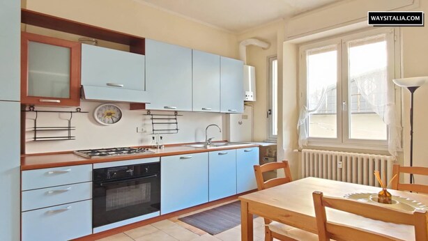 A compact kitchen with light blue cabinets, a wooden countertop, and a dining area with a table and chairs. - De Angeli District Apartment 2 (Milano)