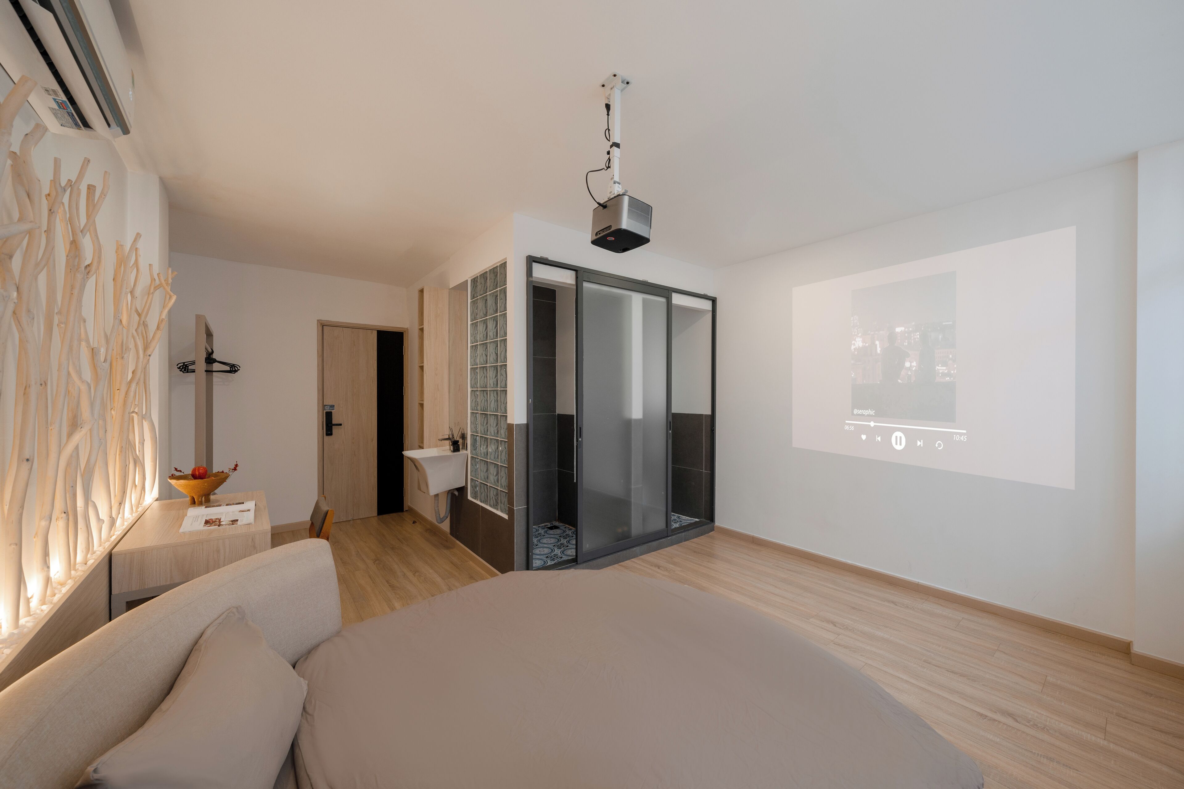 Un dormitorio moderno con una cama grande, una mesita de noche con fruteros, un proyector integrado en la pared y un armario con puerta corredera de cristal.
