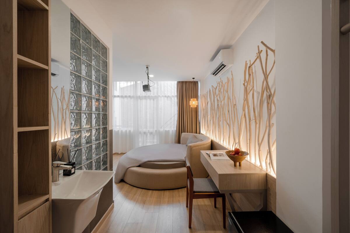 Ein modernes Schlafzimmer mit einem bogenförmigen Bett, einem Holztisch, einem weißen Waschbecken und einem großen Fenster mit transparenten Vorhängen.