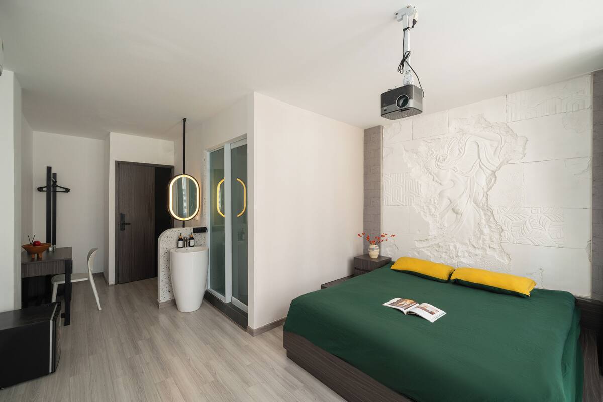 Ein modernes Hotelzimmer mit einem grünen Bett, einer weißen Wand mit einer strukturierten Karte, einem kleinen Nachttisch mit einer Vase und einem großen Spiegel mit rundem Rahmen.