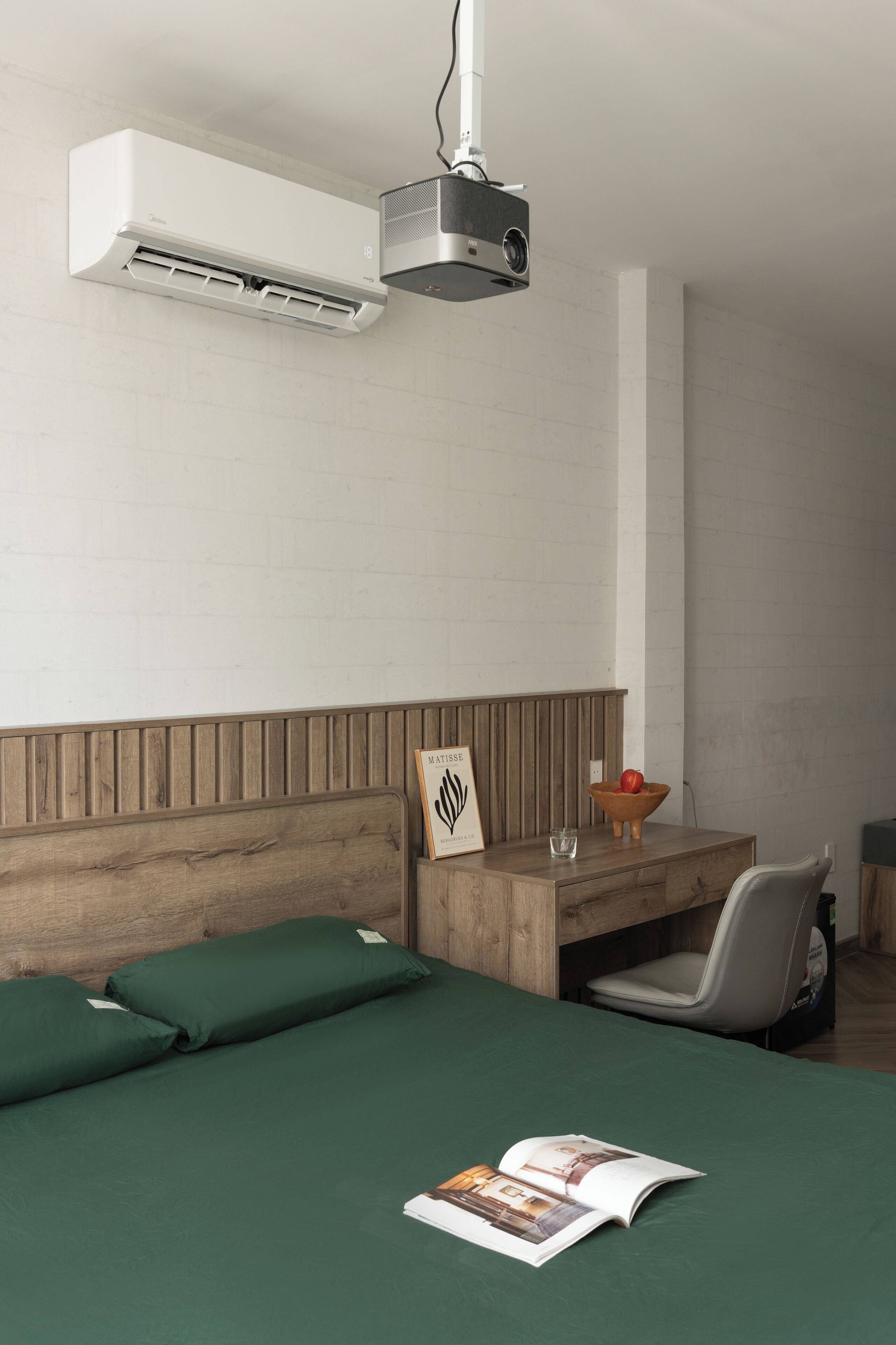 Una moderna camera d'albergo con testiera in legno, un letto con biancheria verde, una scrivania con sedia, un proiettore e un quadro appeso al muro.