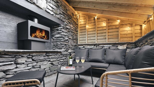 A cozy outdoor seating area with a stone wall, a built-in fireplace, and a cushioned sofa. - Ferry View Cottage in Bowness-on-windermere (Windermere)