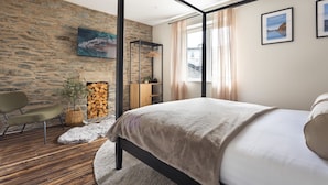 A modern bedroom with a stone wall, a bed with a beige blanket, a wooden chair, a hanging plant, and a fireplace. - Ferry View Cottage in Bowness-on-windermere (Windermere)