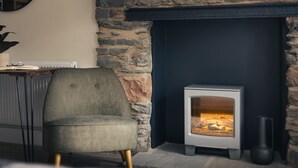 A cozy room with a stone fireplace, a comfortable armchair, and a modern wood-burning stove. - Ferry View Cottage in Bowness-on-windermere (Windermere)
