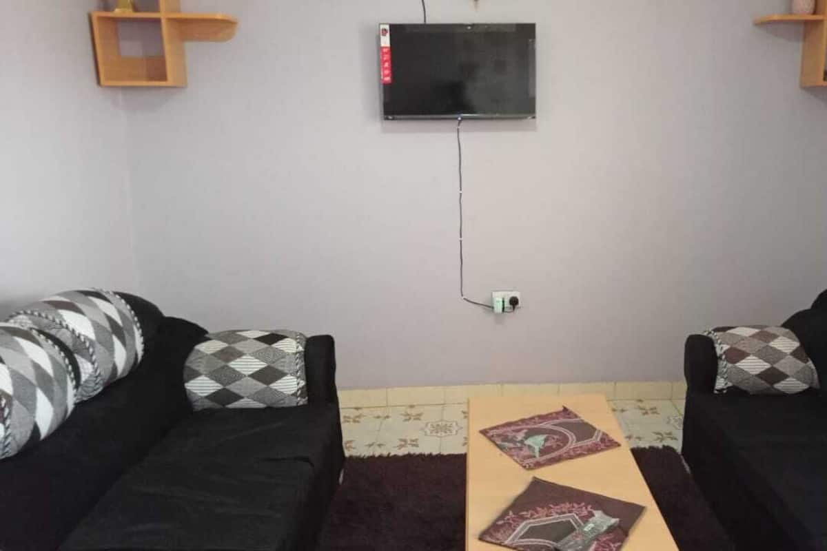 Una sala de estar con dos sofás negros, cojines estampados, un televisor de pantalla plana montado en la pared y un pequeño estante de madera en la pared.