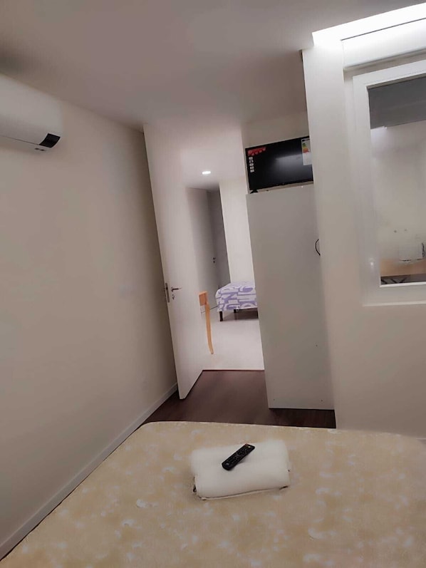 A narrow hallway with a bed in the background, a TV mounted on the wall, and a remote control on the bed. - Casa Augusta 4 (Braga)