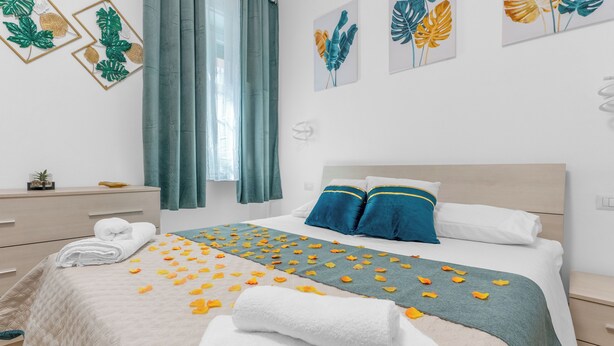 Ein ordentlich bezogenes Bett mit einer grün-gelben Decke, blauen Kissen und einem weißen Kopfteil. Handtücher liegen daneben auf dem Bett und ein kleiner Tisch mit einer Pflanze steht bereit.