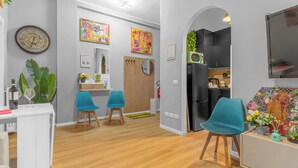 Ein modernes Wohnzimmer mit Holzboden, einem weißen Schreibtisch, zwei blauen Stühlen und einem durch einen Bogen sichtbaren Küchenbereich.