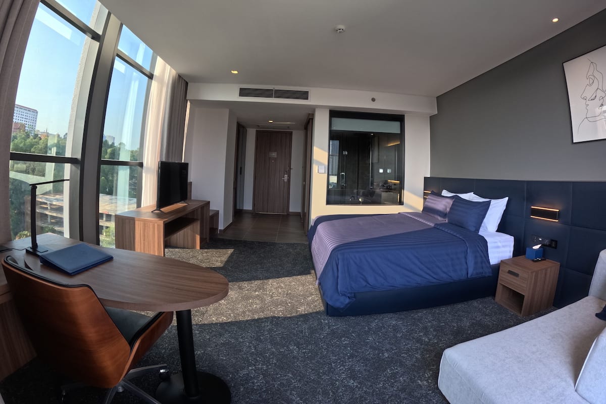 Ein modernes Hotelzimmer mit einem großen Bett, einem Schreibtisch, einem Sessel, einem Fernseher und Blick auf die Stadt durch große Fenster.