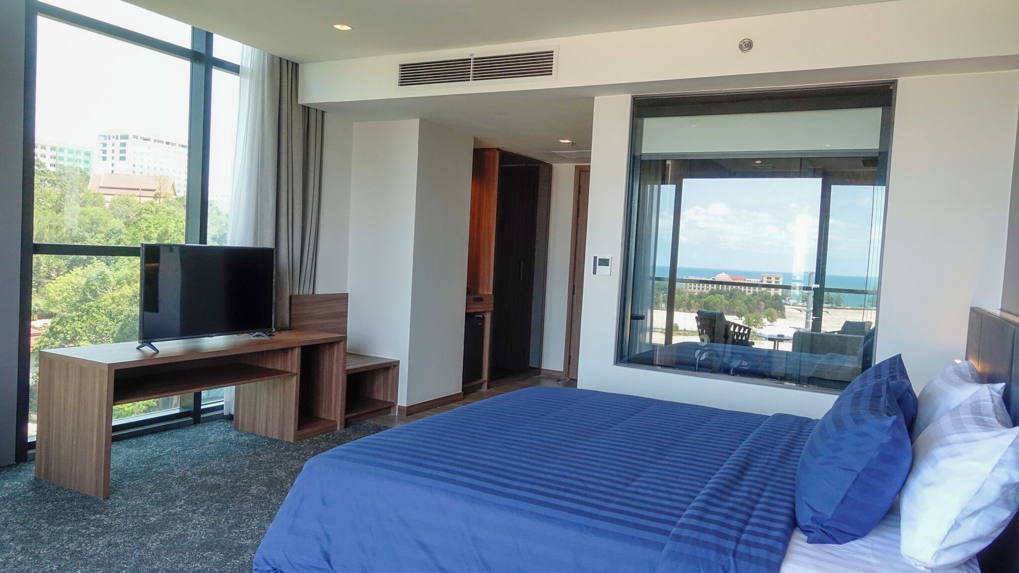 Habitación de hotel con una cama grande, un televisor sobre un escritorio de madera y un balcón con vistas a la ciudad.