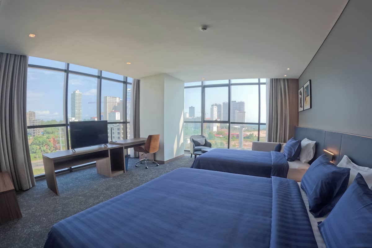 Ein Hotelzimmer mit zwei Betten, einem Schreibtisch, einem Sessel, einem Fernseher und großen Fenstern mit Blick auf die Stadt.