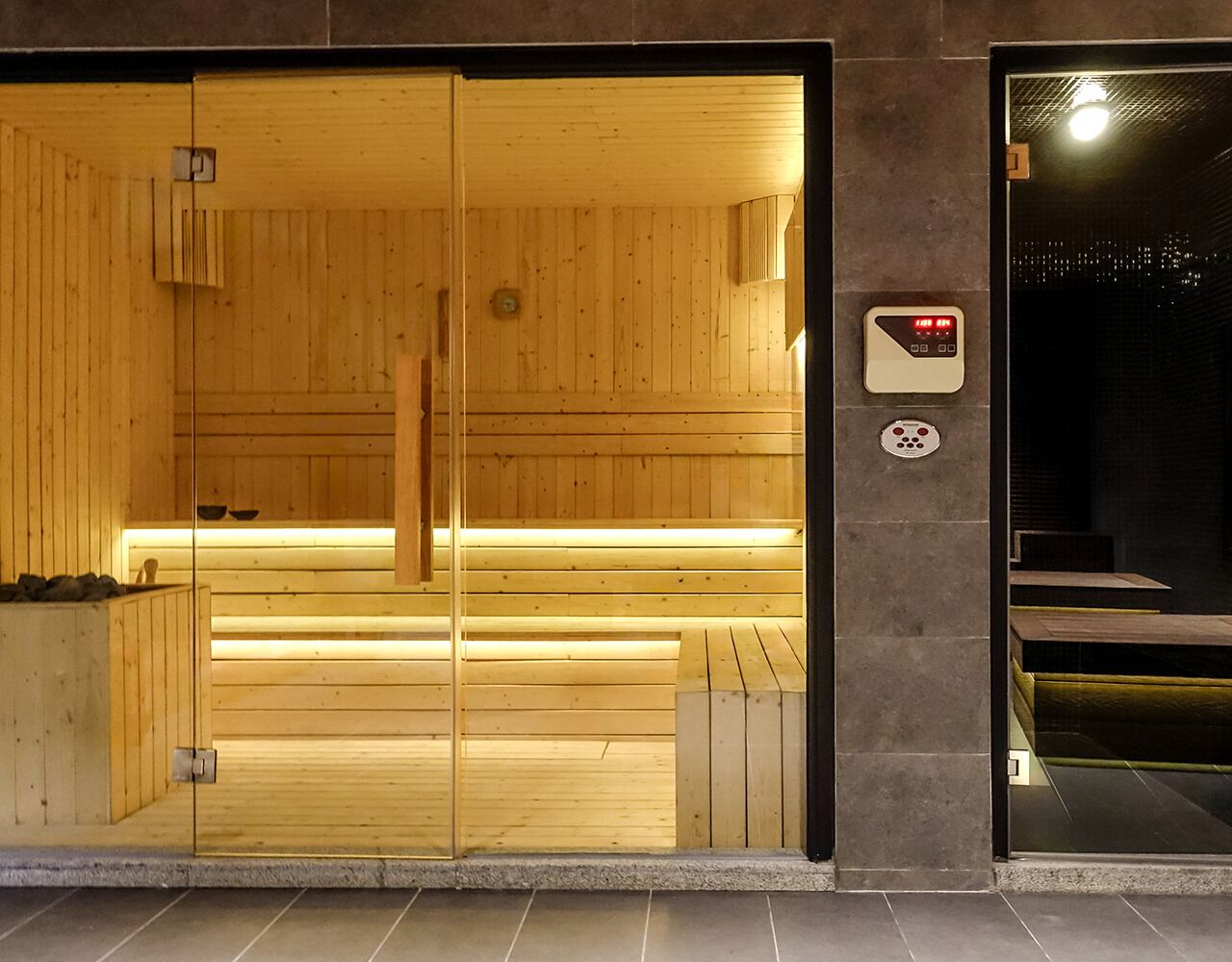 A sauna with wooden benches and a glass door, a control panel, and a tiled floor.