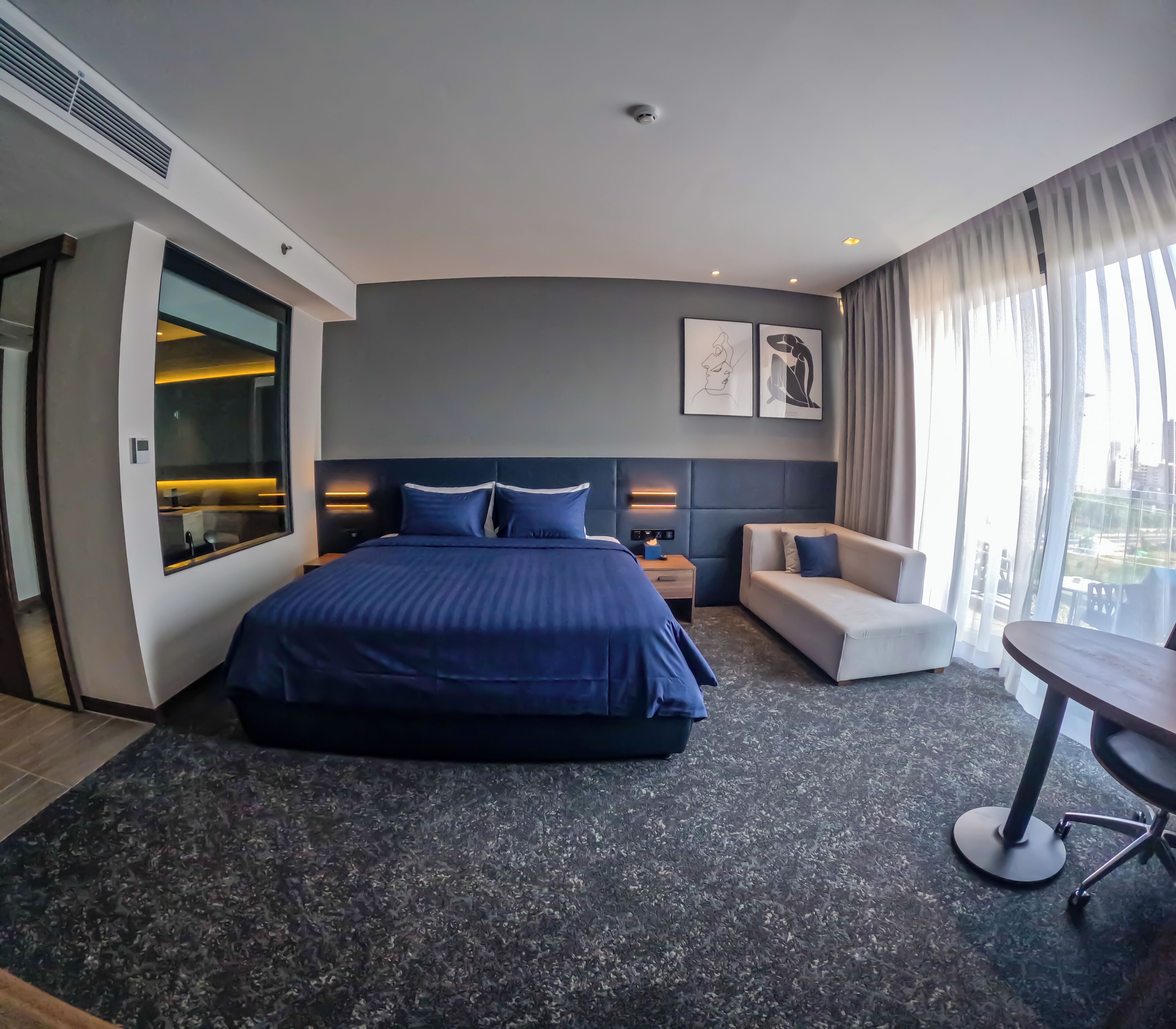 A modern hotel room with a large bed, a seating area with a sofa and armchair, a small table, and a large window with curtains.